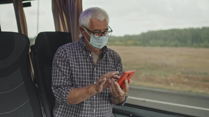 Medium shot of Caucasian senior man in casual checkered shirt and protective face mask using smartphone while sitting by window during bus ride - Powered by Adobe