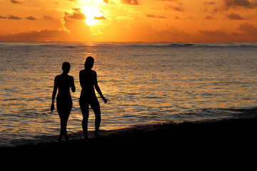 Silhouettes of two girls in bikini walking by the sand on sea beach. Sunset on a coast, travel and holiday concept