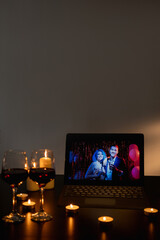 Romantic party. Online greeting. Virtual friends. Two glasses of wine on festive candles decorated table with friendly congrats couple on laptop dark room interior.