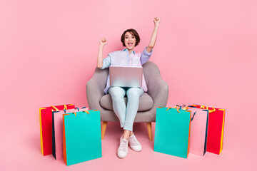 Full length body size photo woman sitting with packages gesturing like winner with laptop isolated pastel pink color background