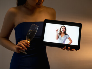Christmas greeting. Online party. Video conference. Unrecognizable woman holding glass champagne tablet computer with happy lady congratulation dark room interior.