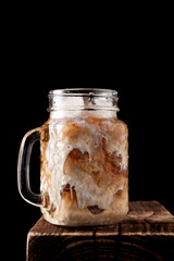 Iced coffee with ice in transparent misted cup on wooden board isolated on black background. Selective focus, copy space