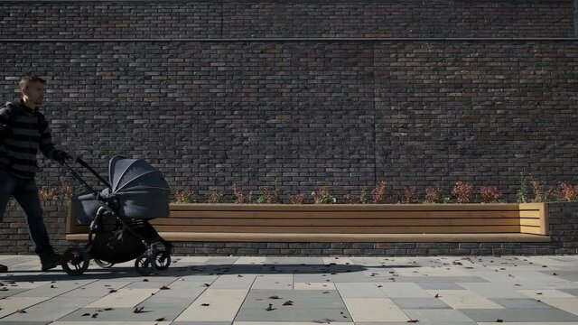 Millennial man in maternity leave with pram and child. Father and his little son walking with newborn baby stroller on the city near brick wall.