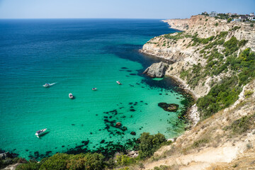Fototapeta na wymiar Boats and yachts in the crystal clear azure sea on a sunny day. Cape Fiolent in Sevastopol. The concept of an ideal place for summer travel and relaxation