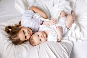 little girl and baby in white clothes are lying in bed on white bed linen. brother and sister bask...