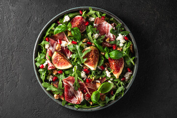 Salad with figs, prosciutto ham, arugula, goat cheese, nuts and pomegranate seeds on black background. top view - obrazy, fototapety, plakaty