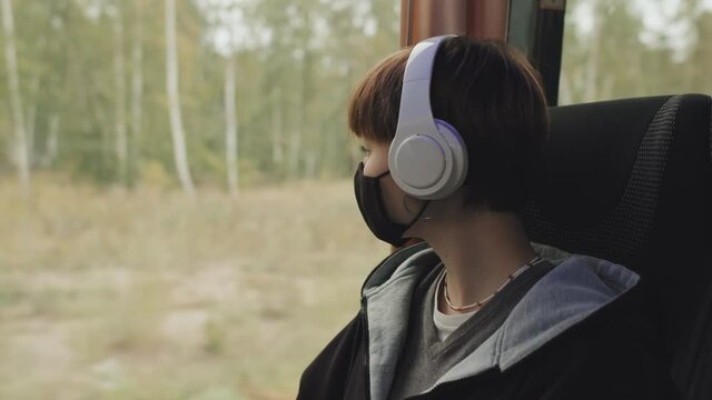 Medium Close-up Of Young Student Girl In Face Mask And Casualwear Listening To Music In Headphones Sitting By Window Alone During Bus Ride