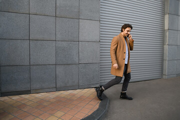 Smiling man in coat talking on smartphone while walking near building 