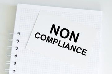 NON COMPLIANCE inscription on a white card against the background of an empty notebook on the table