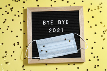 black letter board with text BYE BYE 2021 with small stars and mask on yellow background.