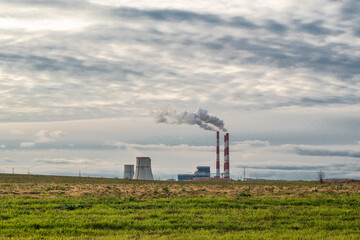 Fototapeta na wymiar Green field against the background of clouds and blue sky, behind it a thermal power plant with smoking chimneys