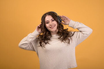 Happy caucasian brunette girl listening to music with headphones, enjoying modern melody, wearing a jumper, isolated on orange studio background.