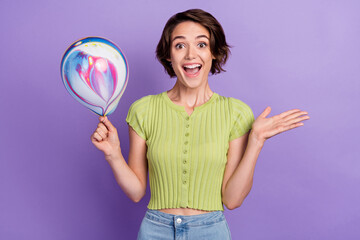 Photo of impressed beautiful young lady wear green t-shirt smiling holding colorful balloon isolated violet color background