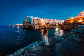 Young woman on the rocks in front of the Apulian village of Polignano a Mare at sunset (blue hour)