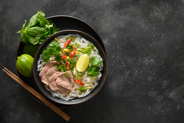 Pho Bo Soup with beef, rice noodles, ginger, lime, chili pepper in bowl on black background. View from above. Copy space. Vietnamese and Asian cuisine.