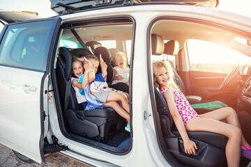 Group of four cute adorable little happy caucasian children enjoy having fun sit in minivan going to sea beach road trip on hot summer day. Kids in swimsuit in car with side open door against sun - obrazy, fototapety, plakaty