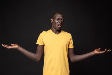 Displeased perplexed young african american man guy in yellow t-shirt posing isolated on black wall background studio portrait. People emotions lifestyle concept. Mock up copy space. Spreading hands.