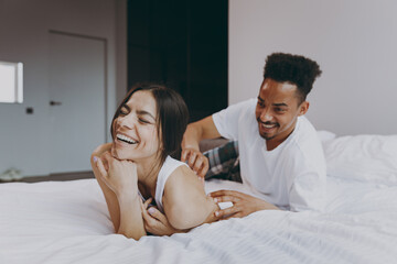 Laughing young couple two family in casual white clothes lying in bed on stomach man tickling woman...