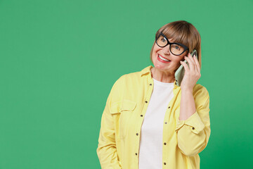 Elderly smiling happy woman 50s in glasses yellow shirt talk speak on mobile cell phone conducting pleasant conversation isolated on plain green background studio portrait People lifestyle concept