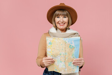 Traveler tourist smiling happy marvelous fun mature elderly senior lady woman 55 years old wears brown shirt hat scarf hold examine map isolated on plain pastel light pink background studio portrait.