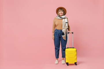 Full size body length traveler tourist vivid mature elderly senior woman 55 years old wears casual clothes hat scarf hold suitcase bag isolated on plain pastel light pink background studio portrait.