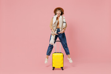 Full size body length traveler tourist mature elderly senior lady woman 55 years old wear casual clothes hat scarf hold suitcase bag jump isolated plain pastel light pink background studio portrait
