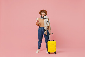 Full size traveler tourist mature elderly lady woman 55 years old wears casual clothes hat scarf hold suitcase bag use mobile cell phone isolated on plain pastel light pink background studio portrait.