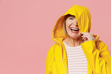 Happy mature elderly lady woman 55 years old wear yellow waterproof raincoat hide cover face with hood isolated plain pastel light pink background studio portrait. Outdoors wet fall weather concept