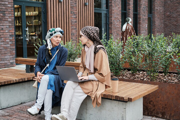 Fototapeta na wymiar Young muslim woman sitting on the bench typing on laptop and discussing something together with other woman outdoors
