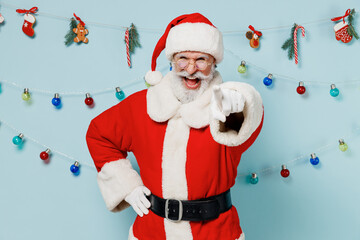 Old laughing bearded Santa Claus man 50s wears Christmas hat red suit point index finger camera on you isolated on plain blue background studio. Happy New Year 2022 celebration merry ho x-mas concept.