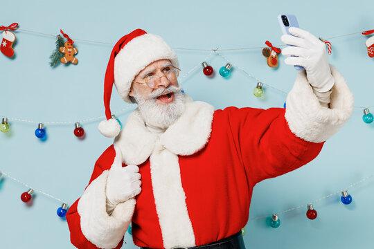 Old bearded Santa Claus man 50s wears Christmas hat red suit doing selfie shot on mobile cell phone show thumb up isolated on plain blue background studio. Happy New Year 2022 merry ho x-mas concept.