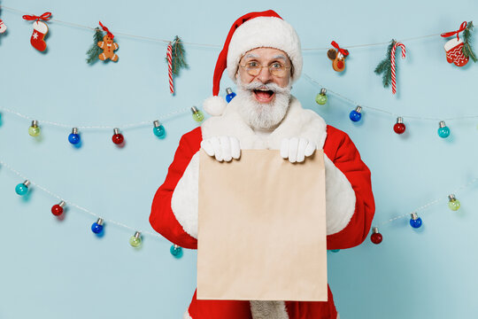 Old Santa Claus man 50s in Christmas hat red suit giving brown craft bag for takeaway mock up with food products isolated on plain blue background studio Happy New Year 2022 merry ho x-mas concept.