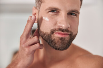 Attractive mature man applies cream onto skin under eye in bathroom