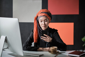 Young muslim businesswoman sitting at her workplace and typing a message on mobile phone she...