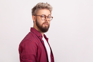 Side view portrait of bearded man in eyeglasses and plaid shirt looking at camera, flirty mood. Indoor studio shot isolated on gray background