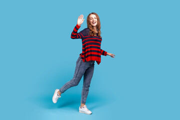 Full length portrait of woman wearing striped casual sweater, standing on one leg and waving hand, saying hello, expressing happiness to meet friend. Indoor studio shot isolated on blue background.