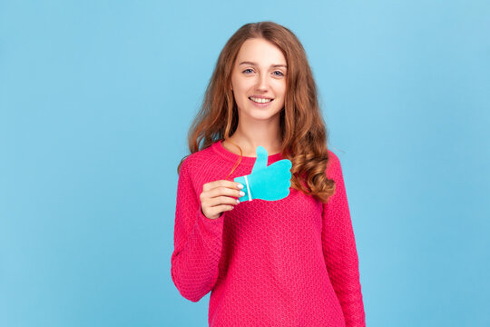 Smiling woman wearing pink pullover, showing thumbs up blue paper sign, liking and recommending posts in social networks, asking to rate. Indoor studio shot isolated on blue background. - Powered by Adobe