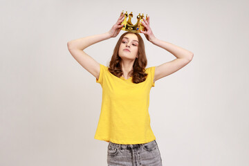 Portrait of selfish brown haired teenage girl in yellow T-shirt holding gold crown over head, having arrogance expression, privileged status. Indoor studio shot isolated on gray background.