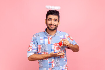 Portrait of attractive bearded man with nimb over head holding small red heart in hands, looking at camera with love, wearing blue casual shirt. Indoor studio shot isolated on pink background.