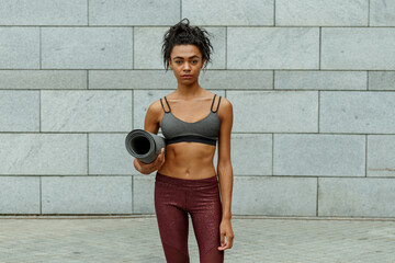 Pretty slim biracial sportswoman in top and leggings holds rolled mat standing against light grey stone wall before training