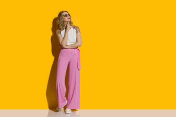 Curious young woman in striped baggy pants is holding hand on chin and looking up.