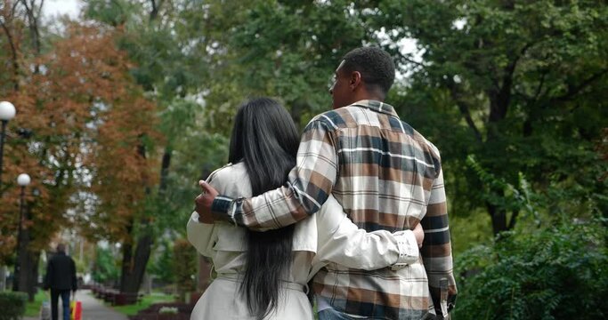 View from the back of a young man in checkered shirt and his girlfriend in beige trench coat hugging each other while walking on the street of a city park, having fun together at romantic date.