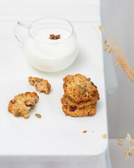 Home made oatmeal cookies with seeds and cranberries. Eco friendly food composition with natural handmade cookies and glass cup of natural milk on a white table with ears of wheat and rye