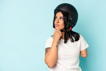 Young caucasian woman with one arm wearing a motorcycle helmet isolated o blue background looking sideways with doubtful and skeptical expression.