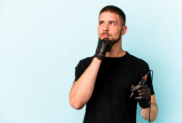 Young caucasian tattoo artist isolated on blue background looking sideways with doubtful and...