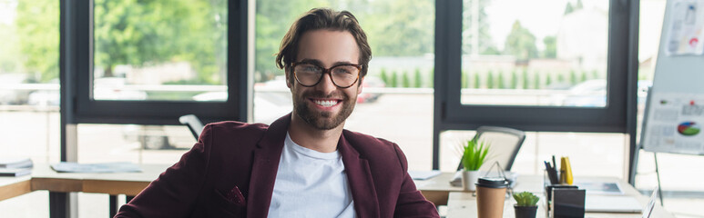 Smiling businessman in eyeglasses looking at camera in office, banner