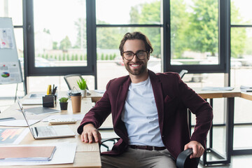 Positive businessman in eyeglasses looking at camera near devices, calculator and papers in office