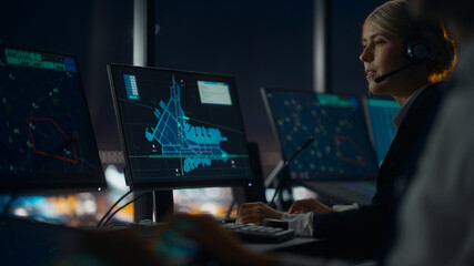 Female Air Traffic Controller with Headset Talk on a Call in Airport Tower at Night. Office Room is Full of Desktop Computer Displays with Navigation Screens, Airplane Flight Radar Data for the Team. © Gorodenkoff