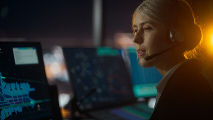 Close Up Female Portrait of Air Traffic Controller with Headset Talk on a Call in Airport Tower at Night. Office Room is Full of Computer Displays with Navigation Screens, Airplane Flight Radar Data.
