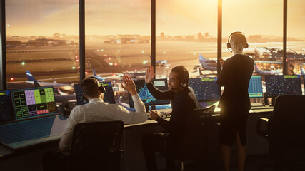 Air Traffic Control Team Working in a Modern Airport Tower at Sunset. Controllers Celebrate Sucessful Landing, Giving High Five to Each Other. Office Room is Full of Navigation Screens, Flight Radars - obrazy, fototapety, plakaty
